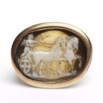 Horizontal oval cameo. White over pale brownish grey layered agate. Depicts Cupid, naked and winged, driving a chariot drawn to right by two horses. Set in a later gold ring.