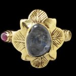 Gold ring, the chased flower-shaped bezel set with a sapphire. The shoulders having settings for stones, one holding a garnet, the other empty